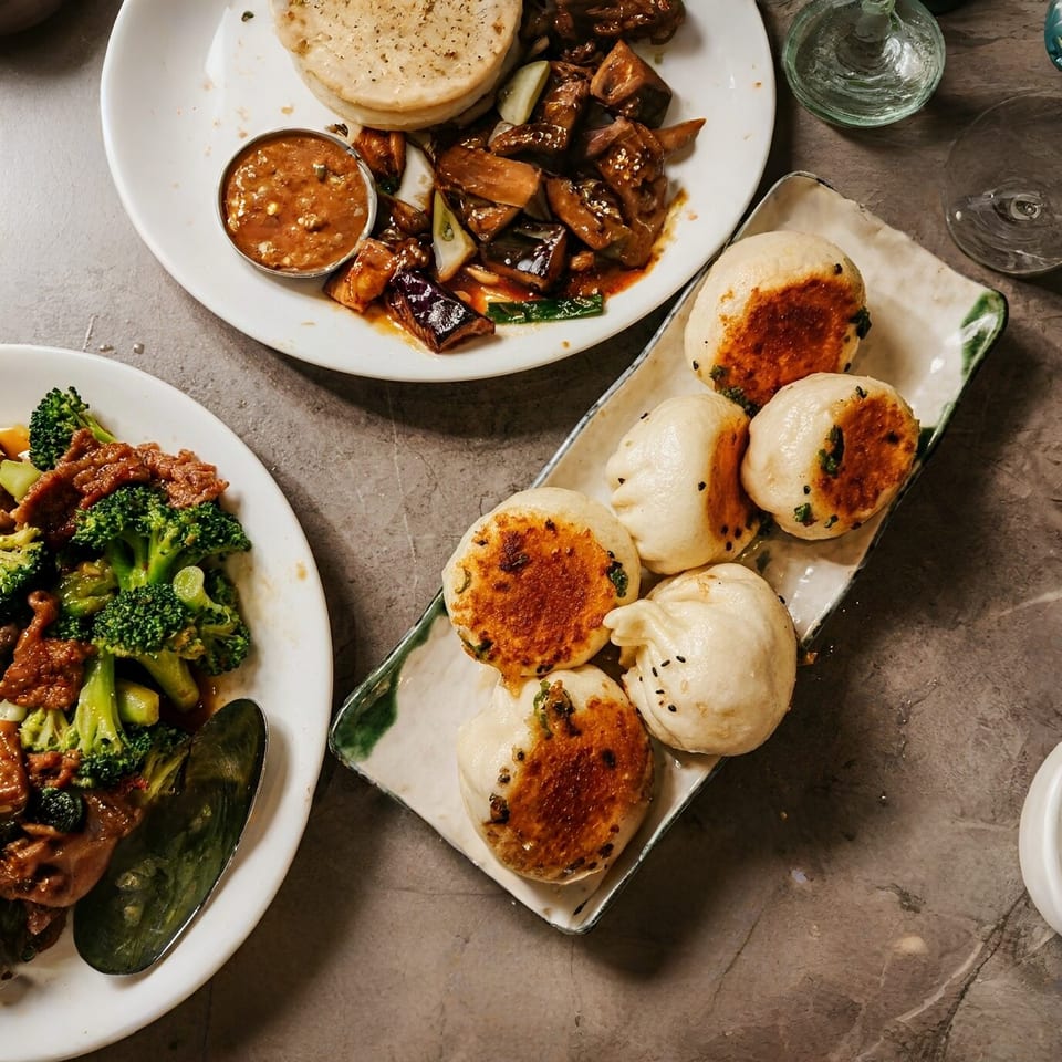 Overhead view of three plates of Asian cuisine, including pan-fried buns, beef and broccoli, and eggplant.