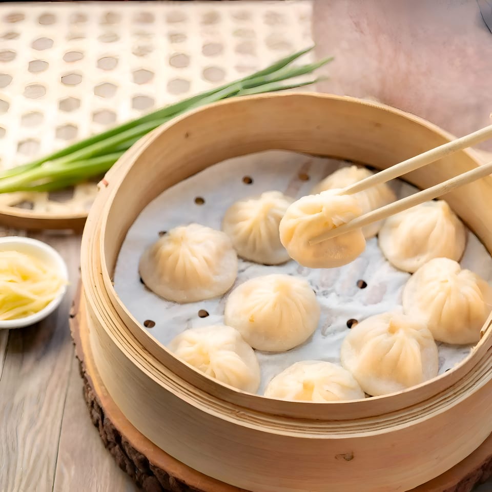 Chopsticks lift a soup dumpling from a bamboo steamer filled with other dumplings, ginger, and scallions.