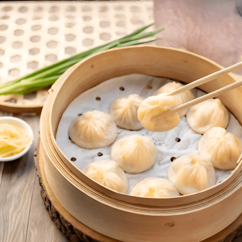 Chopsticks lift a soup dumpling from a bamboo steamer filled with other dumplings, ginger, and scallions.