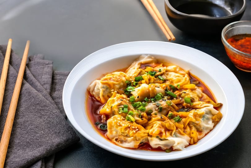 A white bowl filled with dumplings in chili oil, garnished with green onions and sesame seeds.