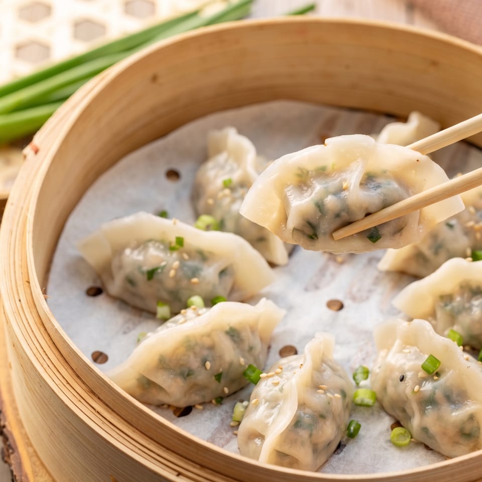 Chopsticks lift a steamed dumpling from a bamboo steamer filled with more dumplings, ginger, and scallions.